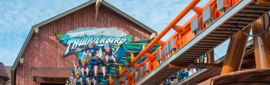 The Thunderbird roller coaster train launches forward on straight, orange track as the Thunderbird logo and station that looks like a barn is behind the train. A man and young boy are seen holding on in the first car, while a young boy in the second row has his hands in the air.