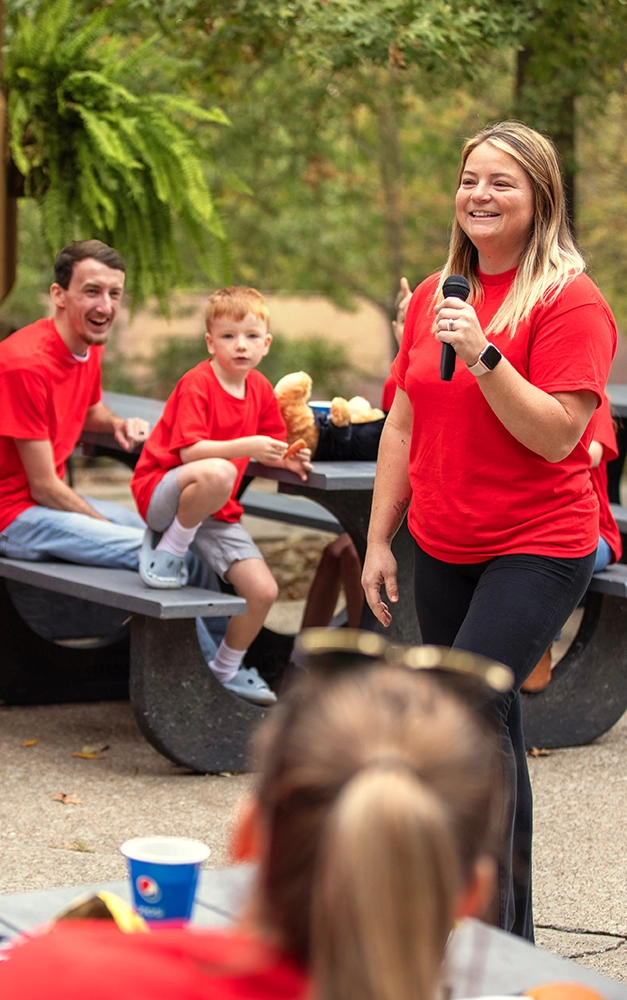 Guests in red shirts sit at picnic tables listening to a woman speaking to them.