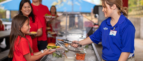 A Team Member serves a young girl in the serving line in the Good Old Days Picnic Grove.